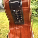 Detail hřístny akustické kytary v hnědé barvě s černým předzesilovačem. Na panelu jsou nápisy VOLUME, BOOST, BASS, MIDD., TRES., PRES. a označení mod…