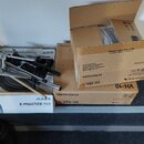 Na fotce jsou tři kartony s hudebním vybavením a kovový stojan. Na spodním bílém kartonu je nápis Alesis E-PRACTICE PAD. Prostřední hnědý karton nese…