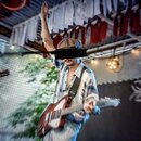 Fotografie zachycuje muže s elektrickou kytarou v hnědém lakovaném provedení. Nástroj má bílý pickguard a světle barevný hmatník. Kvůli rozostření sn…