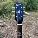 Fotografie zachycuje hlavu a část hmatníku elektrické kytary v černé barvě. Na hlavě je bílý nápis Gibson a pod ním bílé logo ve tvaru kytary. Nástro…