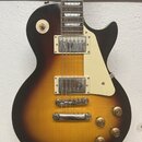 Elektrická kytara s tmelelovým korpusem v barevném přechodu od žluté k tmavě hnědé. Nástroj má dva humbuckery s kovovými kryty, krémový pickguard a č…