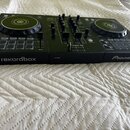 Černý DJ kontrolér se dvěma jog-koly a centrálním mixážním sekcí s knoflíky a posuvníky. Na přední hraně jsou nápisy rekordbox, PHONES a Pioneer DJ.…