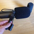 Černý kondenzátorový mikrofon s nasazenou černou pěnovou windscreen. Mikrofon je upevněn v černém plastovém držáku, který drží ruka. V pozadí je vidě…