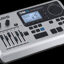 Stříbrný elektronický bicí modul značky ALESIS, model DM10. Na zařízení je nápis HIGH DEFINITION DRUM MODULE WITH DYNAMIC ARTICULATION a displej s te…