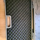 Otevřený přepravní kufr z tmavě šedého plastu. Vnitřek je vyplněn černou akustickou pěnou s vlnovitým profilem, na které jsou vidět prach a jemná vlá…