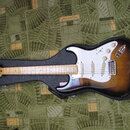 Elektrická kytara v černém tvrdém kufru. Nástroj má dvoubarevný sunburst povrch, bílý pickguard a hmatník ze světlého dřeva. Na hlavě je viditelné lo…