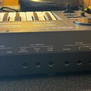 Zadní strana černého klávesového nástroje s logem Roland na horní hraně. Jsou zde viditelné nápisy MEMORY PROTECT (ON OFF ON), TAPE (LOAD, SAVE), EXT…