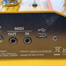 Záběr na zadní panel žlutého hudebního zařízení s černým plastovým krytem. Jsou zde vidět konektory MIDI OUT, MIDI IN a USB port s odpovídajícími náp…
