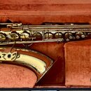 Zlatý saxofon v otevřeném černém kufru s červeným polstrováním. Nástroj je v rozloženém stavu, vedle něj leží samostatně hnedbek. Na těle nástroje ne…