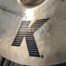 Detailní záběr na povrch kovového činele zlatavé barvy. Uprostřed je vidět velké černé logo ve tvaru písmene „K“ složeného z vodorovných čar, vedle k…