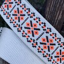 Detail tkaného popruhu v bílé barvě s geometrickým vzorem v hnědé a oranžové barvě. Povrch je strukturovaný, materiál vypadá jako textil. Žádné značk…