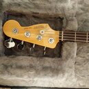 Fotografie zachycuje hlavu basové kytary v barvě světlého dřeva, která leží v polstrovaném kufru. Na hlavě je umístěn nápis JAZZ BASS® a logo Fender.…