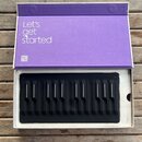 Otevřená fialová krabice s nápisem „Let's get started“ a QR kódem. Uvnitř je v bílém polystyrenovém lůžku umístěn černý MIDI kontrolér s gumovou kláv…
