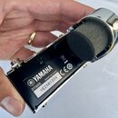 Fotografie zachycuje detail zadní strany černého mikrofonu s chromovaným kroužkem a černou větrovou popsávkou. Na černé ploše je bílý nápis YAMAHA a…