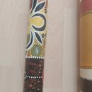 Detailní pohled na povrch didgeridoo s barevným ornamentem. Nástroj je zdoben ručně malovanými vzory v žluté, bílé a červené barvě na tmavém podkladu…