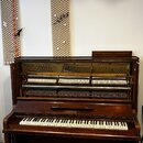 Hnědý dřevěný pianino s otevřeným horním víkem, skrze které je vidět vnitřní mechanismus a struny. Na předním panelu nad klávesnicí je umístěn nápis…