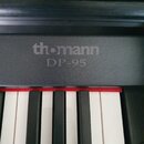 Detail digitálního piana v černé barvě. Na horní části korpusu je šedý nápis th·mann a pod ním označení DP-95. V dolní části jsou vidět bílé a černé…