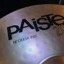 Detailní záběr na kovový činel stříbrné barvy s jemným žebrováním povrchu. Na čineli je černý nápis PAiste a pod ním text 18" CRASH RIDE. Vpravo od l…
