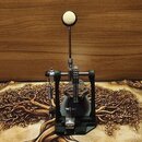 Fotografie zobrazuje bicí pedál v černé barvě s kovovými mechanismy. Na horní části beateru je bílá plstěná hlavička. Nástroj nevykazuje viditelné zn…