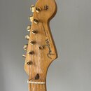 Fotografie zachycuje hlavu elektrické kytary z přírodního dřeva se zlatými ladičkami. Na povrchu je vidět logo Fender a nápis STRATOCASTER s doplňkem…
