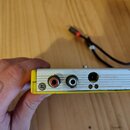 Detail zadní strany žlutého zařízení s hliníkovým povrchem. Jsou vidět dva RCA konektory (červený a bílý) a jeden kulatý napájecí vstup. Povrch vykaz…