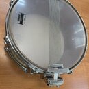Fotografie znázorňuje snare drum (malý táč) s kovovým obručováním a bílým membránou. Na membráně je černý nápis YAMAHA STATIC 115WG HEAD MADE IN U.S.…