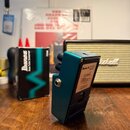 Kytarový efekt pedál v tyrkysové barvě s černým spodním krytem. Na spodní straně je bílý štítek s logem Ibanez a nápisem POWER SUPPLY. Vedle pedálu j…