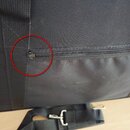 Detailní záběr na černý textilní obal s zipem. Červený kruh zvýrazňuje kovový jezdec zipu. V dolní části je vidět černý popruh s kovovými karabiny.