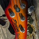 Detail hlavy basové kytary v červeno-oranžovém přechodovém odstínu. Na dřevě je černý nápis „Sterling by MUSIC MAN“ a v kroužku označení „Ray34“. Jso…