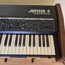 Černý syntetizátor s dřevěnými bočními panely. Na horním panelu je bílý nápis JUPITER-4 COMPUPHONIC a označení sekcí VCO1, NOISE, VCF a VCA. Nástroj…