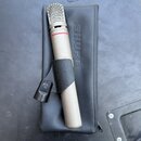 Stříbrný mikrofon s kovovou mřížkou a červeným prstenem pod hlavou. Mikrofon je upevněn v černém držáku a položen na černém pouzdře s vyraženým nápis…