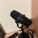Černý dynamický mikrofon s nasazenou pěnovou windsockem, umístěný v držáku na stojanu. Na těle mikrofonu je bílý nápis SHURE a ovládací prstenec s oz…