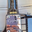 Detail zadní strany krku a spoje s tělem černé elektrické kytary. Na hmatníku je nalepený štítek s nápisem MODEL JR-SC/BK. Kovový kryt s logem Epipho…