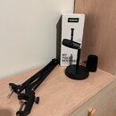 Na světlém dřevěném povrchu je černý mikrofonní rameno a černý stojan s mikrofonem. Vedle nich stojí bílá krabice s nápisy SHURE, MV7 PODCAST MICROPH…