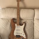 Elektrická kytara s přírodním dřevěným povrchem těla a hmatníkem. Na hlavě je viditelné logo Fender Stratocaster. Bílý pickguard obsahuje tři snímače…