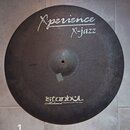 Kulatý činel s tmavým, matným povrchem. Na horní části je bílý nápis „Xperience“ a pod ním „X-jazz“, vlevo od něj je menší nečitelný text. Ve spodní…