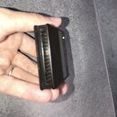 Černý plastový modul s konektorem s řadou zlatých pinů. Na bočním povrchu je viditelný nečitelný nápis. Předmět drží ruka nad šedým povrchem.