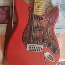 Červená elektrická kytara typu Stratocaster s hnedým dřevěným pickguardem a světle žlutým hmatníkem. Na kobylce je viditelný nápis "Wilkinson". Kytar…