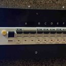 Černý rackový panel s elektroinstalací. Vlevo jsou tři modré digitální displeje zobrazující hodnoty 241V 00.0A (L1), 238V 00.0A (L2) a 241V 00.0A (L3…