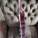 Detailní pohled na elektrickou kytaru z profilu, položenou na hnědém čalouněném křesle. Nástroj má tmavě červený lakovaný povrch s viditnou strukturo…