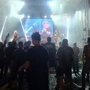 Fotografie zachycuje hudební kapelu vystupující na osvětleném pódiu před publikem. Na scéně jsou vidět hudebníci s nástroji, včetně kytary a bicích,…