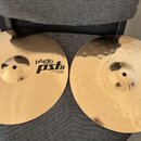 Dvě mosazné činely položené na šedém textilním povrchu. Horní činel má hladký povrch a nápis PAiste PSL 8 14" HI-HAT BOTTOM. Dolní činel má kovaný po…