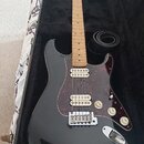 Černá elektrická kytara s tvarem typu Stratocaster, světlým hmatníkem a červeně vzorovaným pickguardem. Nástroj má tři snímače, tři ovládací knofíky…