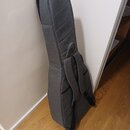 Šedý polstrovaný obal na hudební nástroj s ramenními popruhy. Obal je vyroben z textilie a opřen o bílou zeď na dřevěné podlaze. Na povrchu nejsou vi…