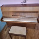 Hnědé dřevěné piano s přiloženou stolečkou s béžovým čalouněním. Na krytu klávesnice je černý nápis Röster. Povrch dřeva vykazuje známky běžného opot…