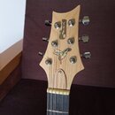 Fotografie zachycuje hlavu světlé dřevěné elektrické kytary s šesti chromovanými ladičkami. Na dřevě je umístěn kovový nápis „Bcg Music“ s motivem no…