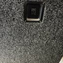 Detailní záběr na část povrchu reproduktorového boxu potaženého šedým kobercovým materiálem. V horní části je umístěn čtvercový plastový panel s jedn…