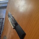 Detailní pohled na kobylku akustické kytary z tmavého dřeva na světle hnědé desce s viditnou sletou. V bílém kostěném nebo plastovém sedle jsou upevn…