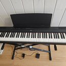 Černá digitální pianola na černém X-stánovku s připozeným pedálem. Na horní liště je uprostřed bílý nápis YAMAHA a vpravo označení modelu P-115. Nást…