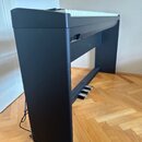 Černý digitální piano v doprovodném stojanu s třemi stříbrnými pedály. Nástroj má bílé klávesy a matný povrch korpusu. Na viditelných částech nejsou…